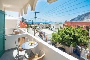 A balcony with wicker chairs, a small table with fruit, and a view of a coastal town with buildings and a mountain in the background. - Ifestia (Santorini)