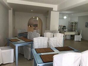 A dining area with tables and chairs, a kitchen in the background, and a hallway leading to another room. - Ifestia (Santorini)