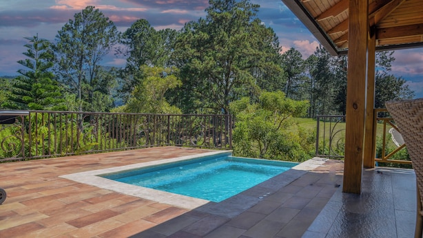 A swimming pool with a wooden deck and a metal fence, surrounded by trees and a view of the landscape. - Vista Del Campo Country Club & Villas Srl (Jarabacoa)
