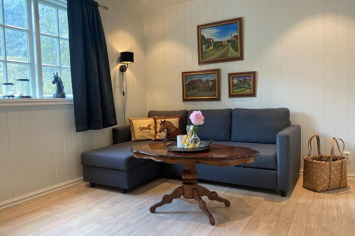 A living room with a grey sofa, a wooden coffee table with a vase and tray, a lamp, and framed paintings on the wall.