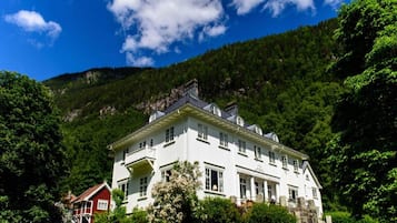 A large white building with multiple windows and a balcony, surrounded by lush greenery and a mountainous backdrop.