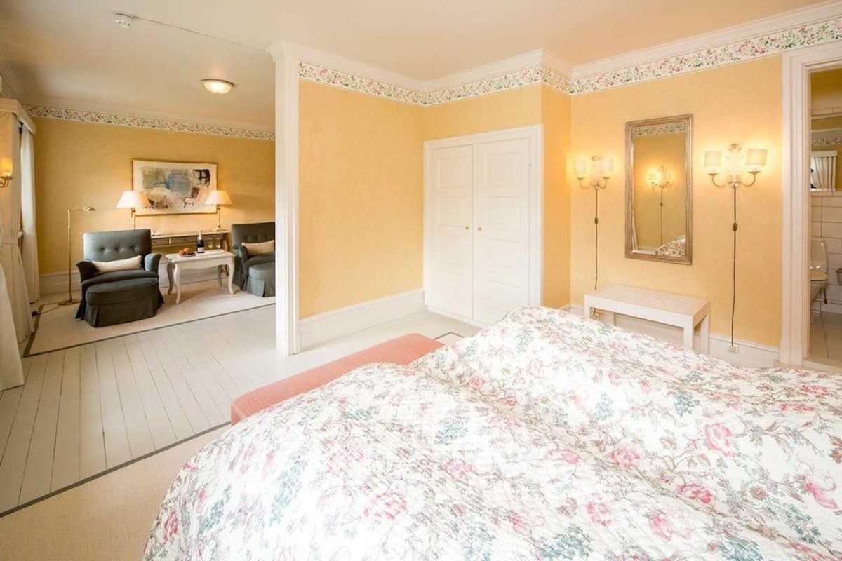 A bedroom with a bed, a desk, two chairs, a wardrobe, and a bathroom visible through an open door.
