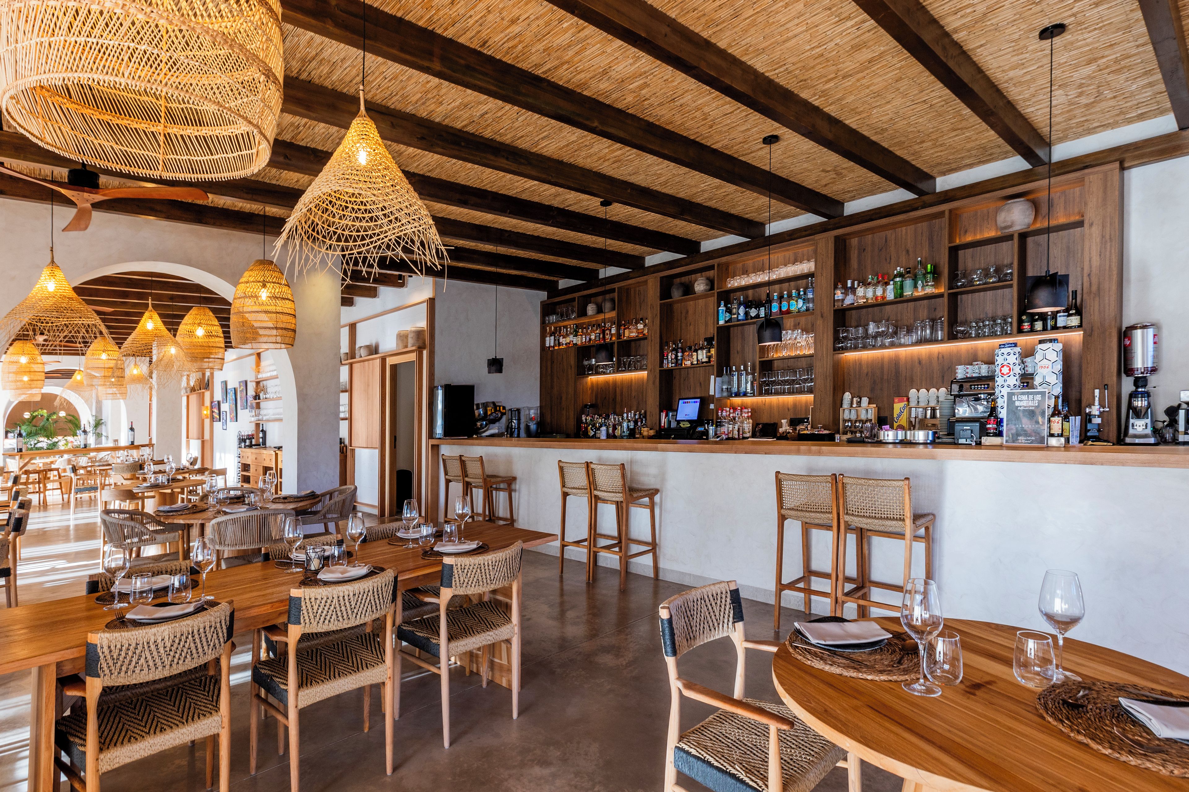 A restaurant with wooden tables and chairs, a bar area with shelves of bottles, and hanging wicker light fixtures.