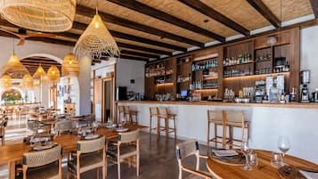 A restaurant with wooden tables and chairs, a bar area with shelves of bottles, and hanging wicker light fixtures.