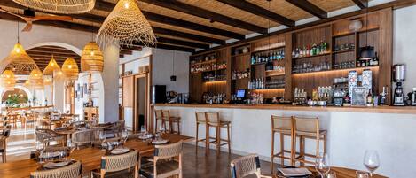 A restaurant with wooden tables and chairs, a bar area with shelves of bottles, and hanging wicker light fixtures.