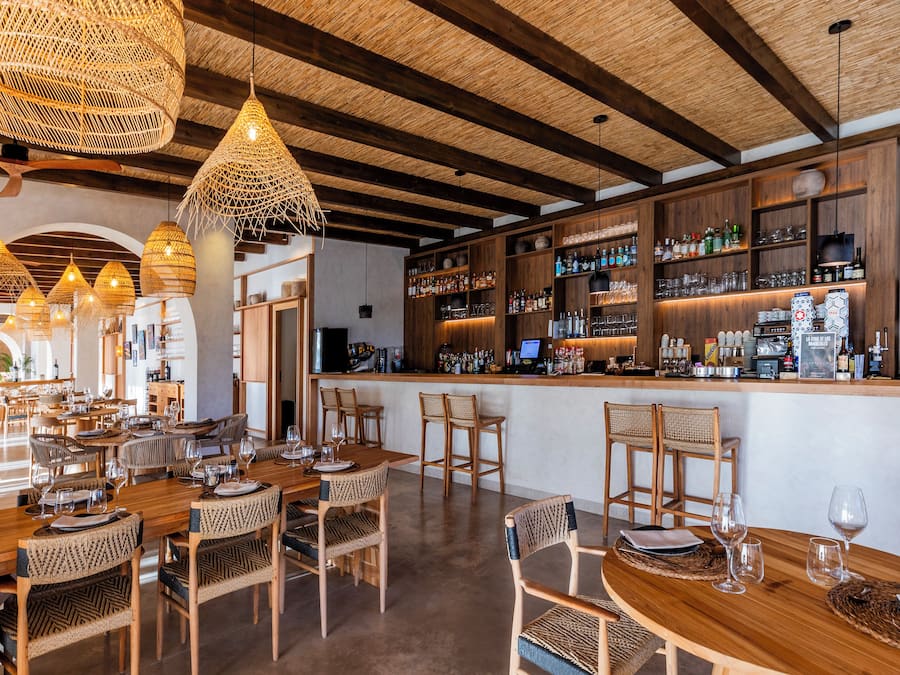 Un restaurante con mesas y sillas de madera, una zona de bar con estanterías llenas de botellas, y lámparas colgantes de mimbre.