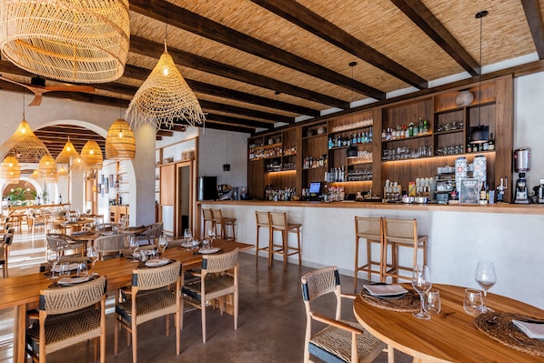Un restaurante con mesas y sillas de madera, una zona de bar con estanterías llenas de botellas, y lámparas colgantes de mimbre.