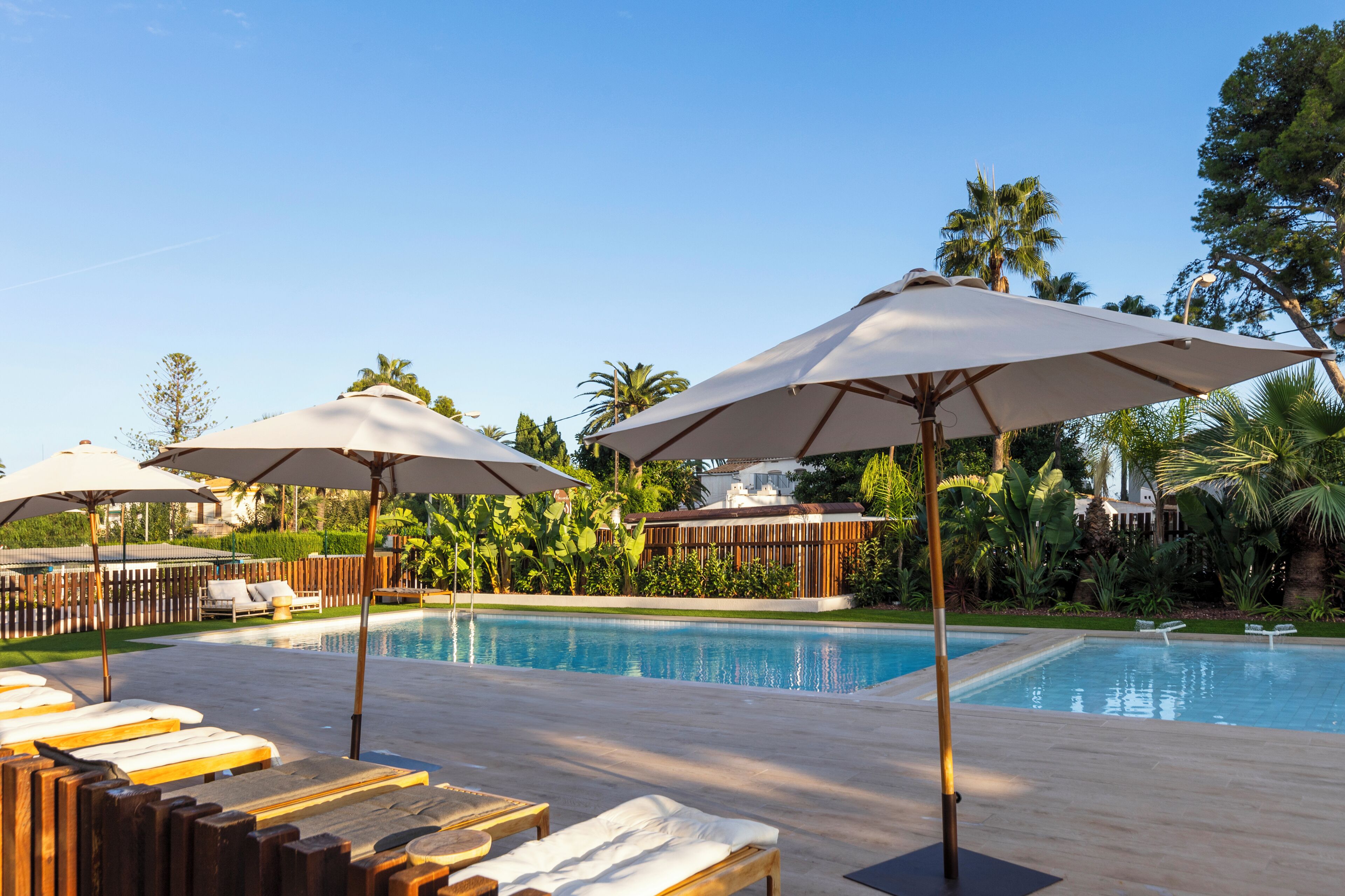 Zona de piscina amb gandules i ombrel·les, envoltada de vegetació i palmeres.