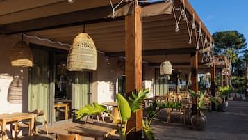 Un espacio al aire libre con mesas y sillas de madera, plantas en macetas y lámparas de mimbre colgantes.