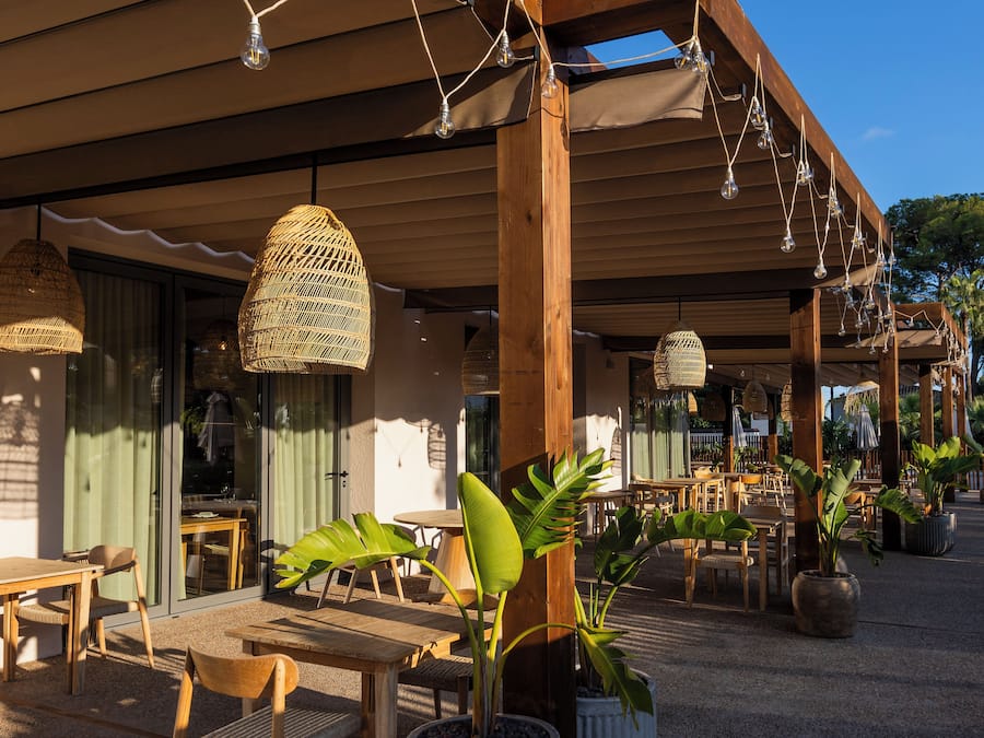 An outdoor seating area with wooden tables and chairs, potted plants, and hanging wicker lamps.