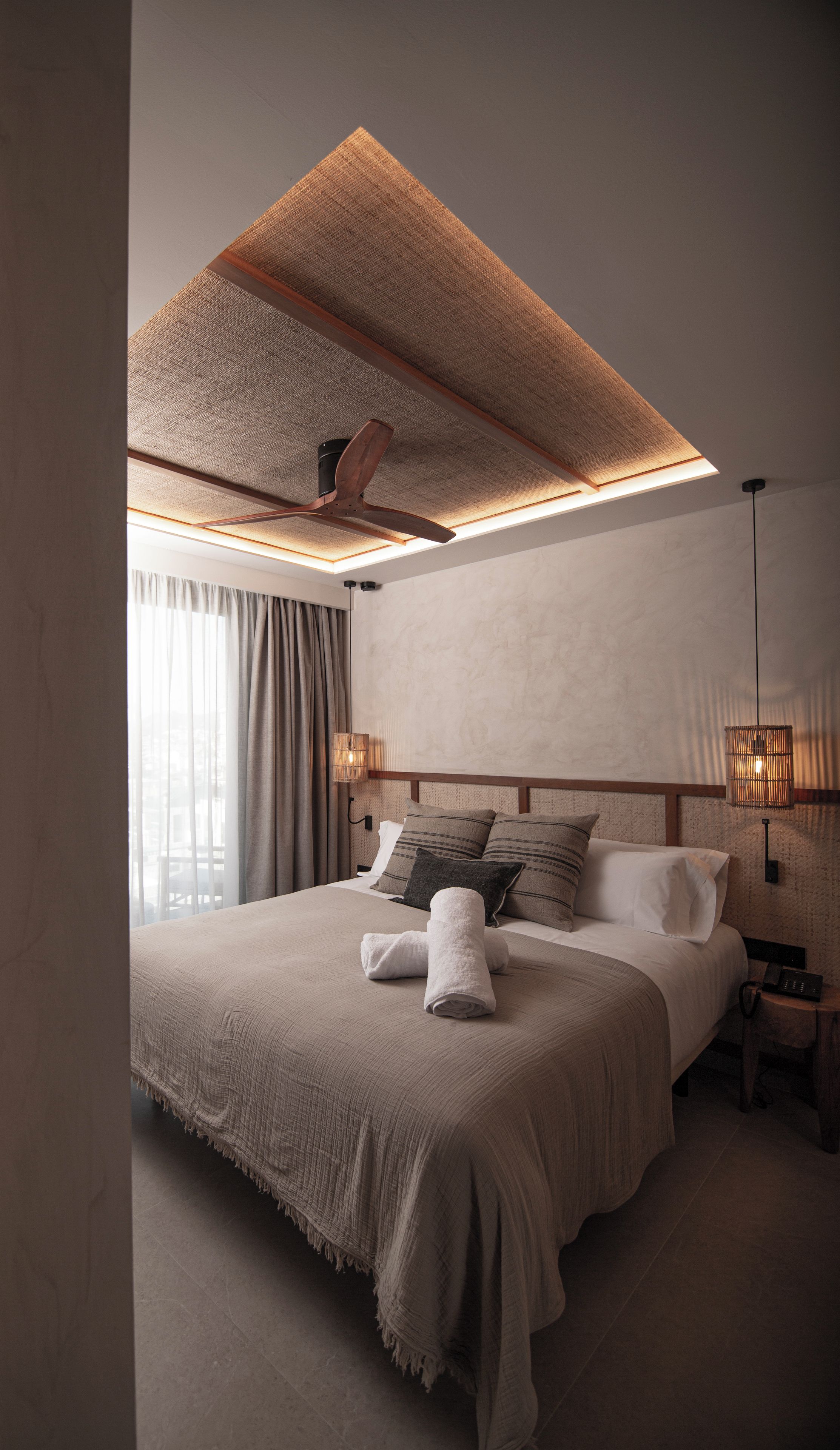 Habitació d'hotel moderna amb un llit gran, un ventilador de sostre, una taula de nit amb una làmpada i vista a l'exterior per la finestra.