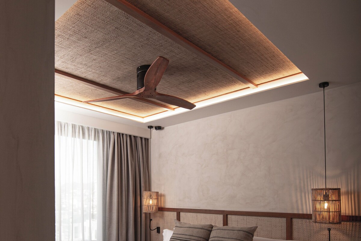 A modern hotel room with a large bed, a ceiling fan, a bedside table with a lamp, and a view of the outdoors through the window.