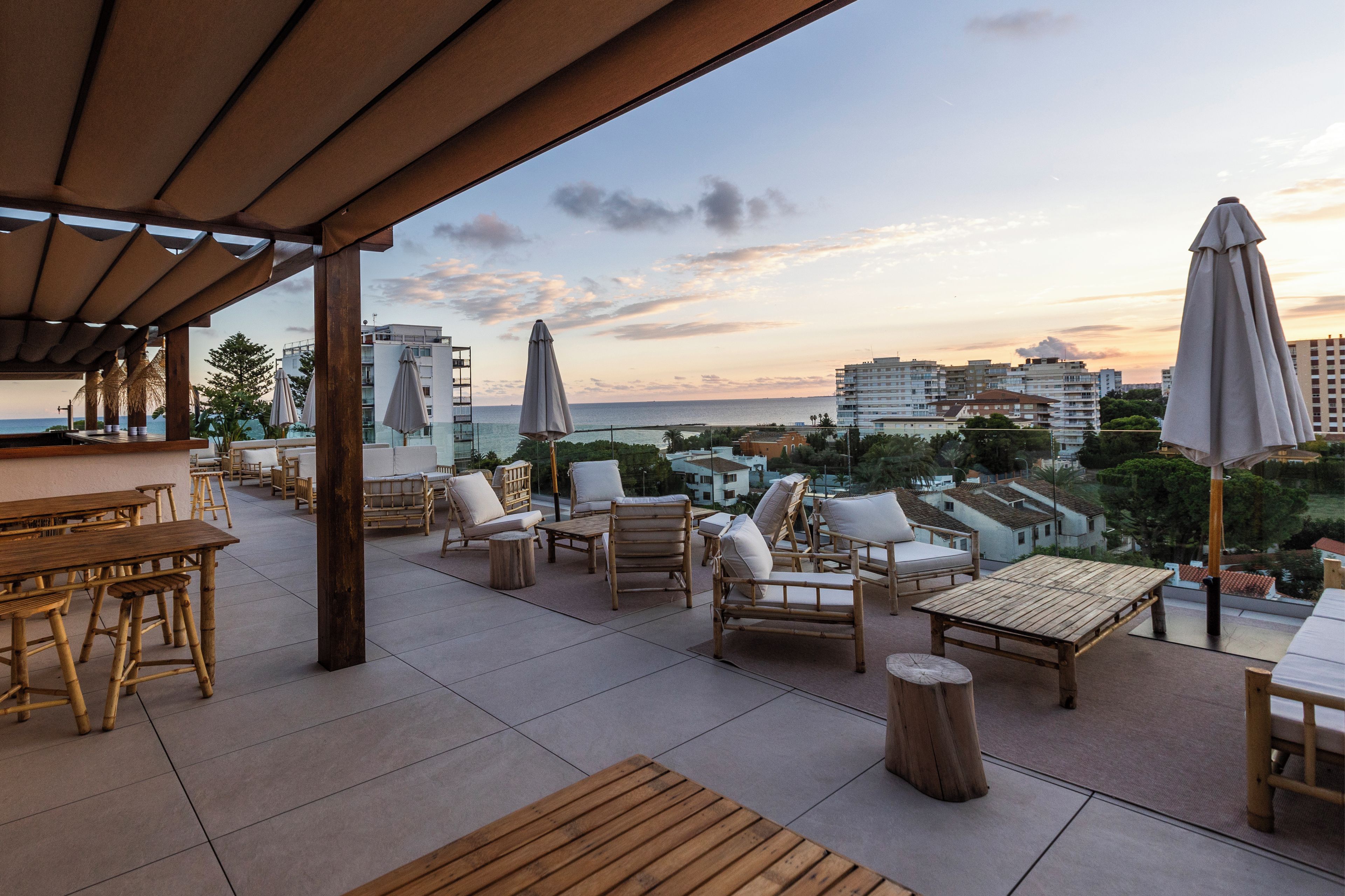 Un área de estar al aire libre con mobiliario de madera, sombrillas y vistas al mar y edificios.