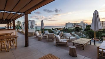Un área de estar al aire libre con mobiliario de madera, sombrillas y vistas al mar y edificios.