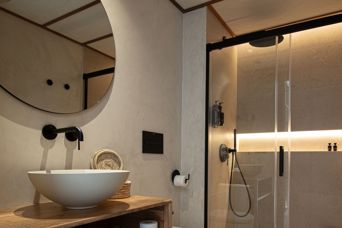 Un baño moderno con un espejo ovalado grande, un tocador de madera con lavabo, un inodoro y una ducha con mampara de cristal.
