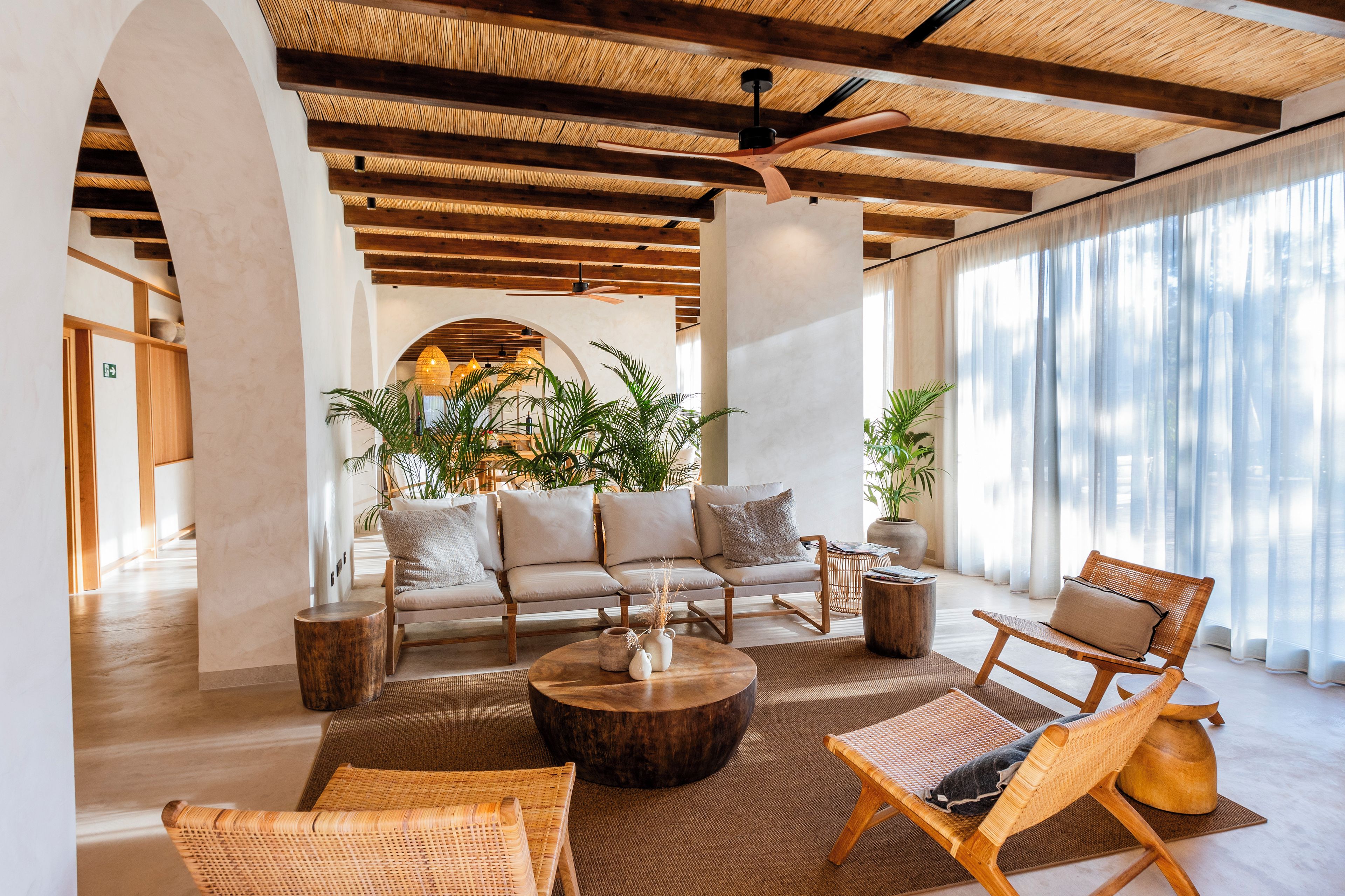 A spacious lounge area with a sofa set, wicker chairs, a coffee table, and large windows with sheer curtains.
