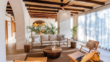 A spacious lounge area with a sofa set, wicker chairs, a coffee table, and large windows with sheer curtains.