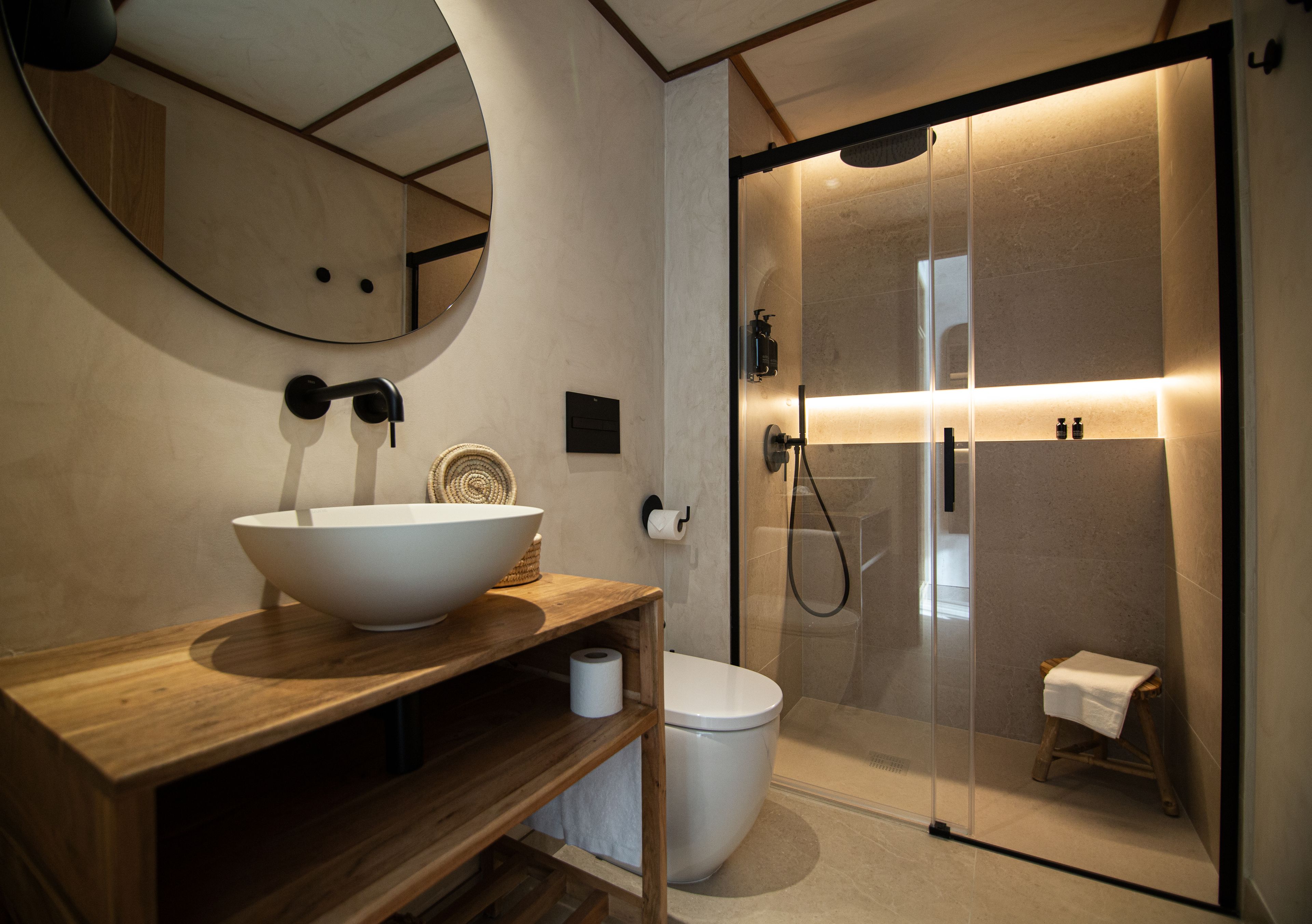 Un baño moderno con tocador de madera, un espejo grande, un lavabo blanco, un inodoro y zona de ducha con mampara de cristal.