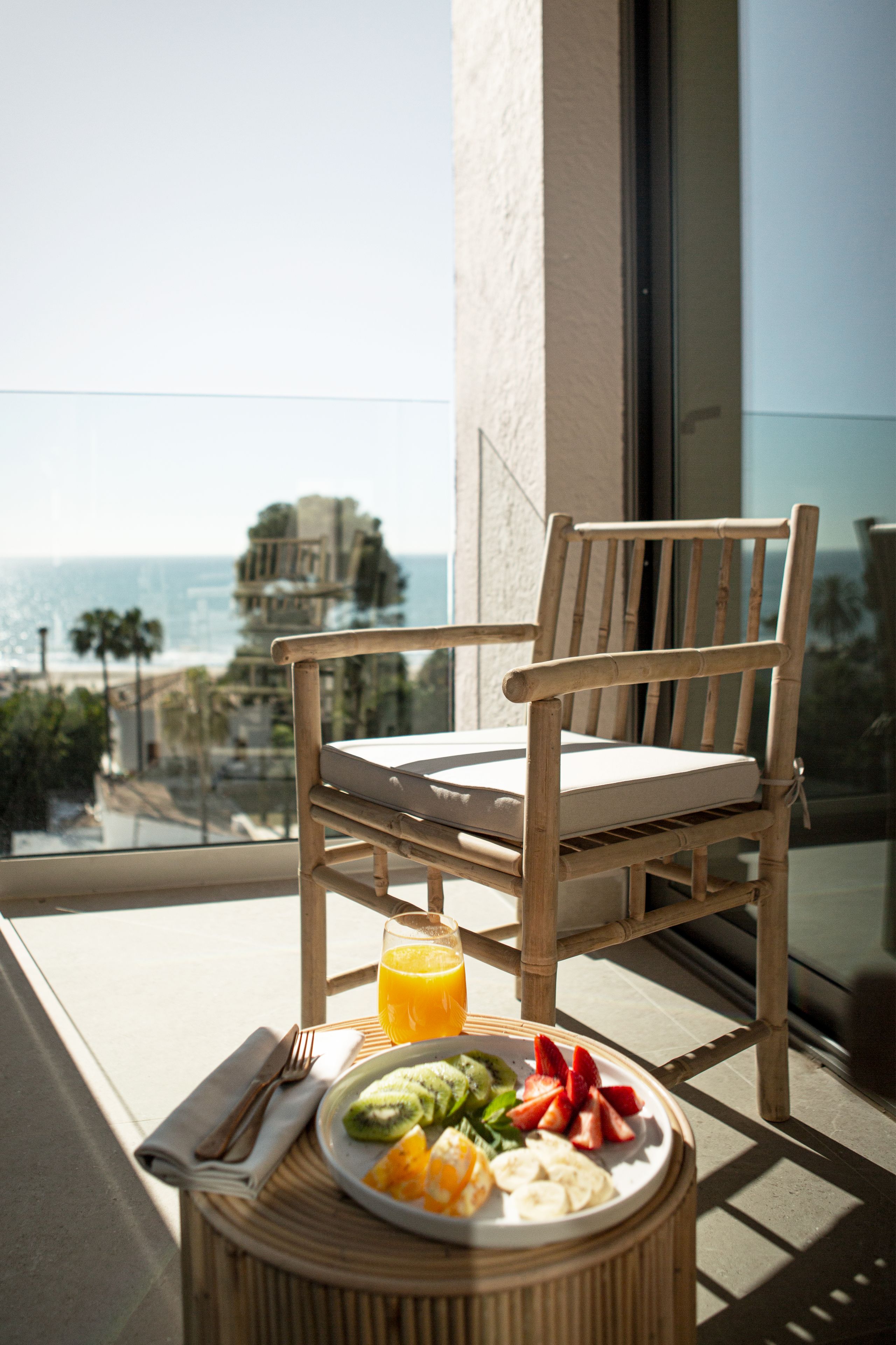 Una silla de madera con cojín en un balcón, una mesa, un plato de fruta y un vaso de jugo de naranja.