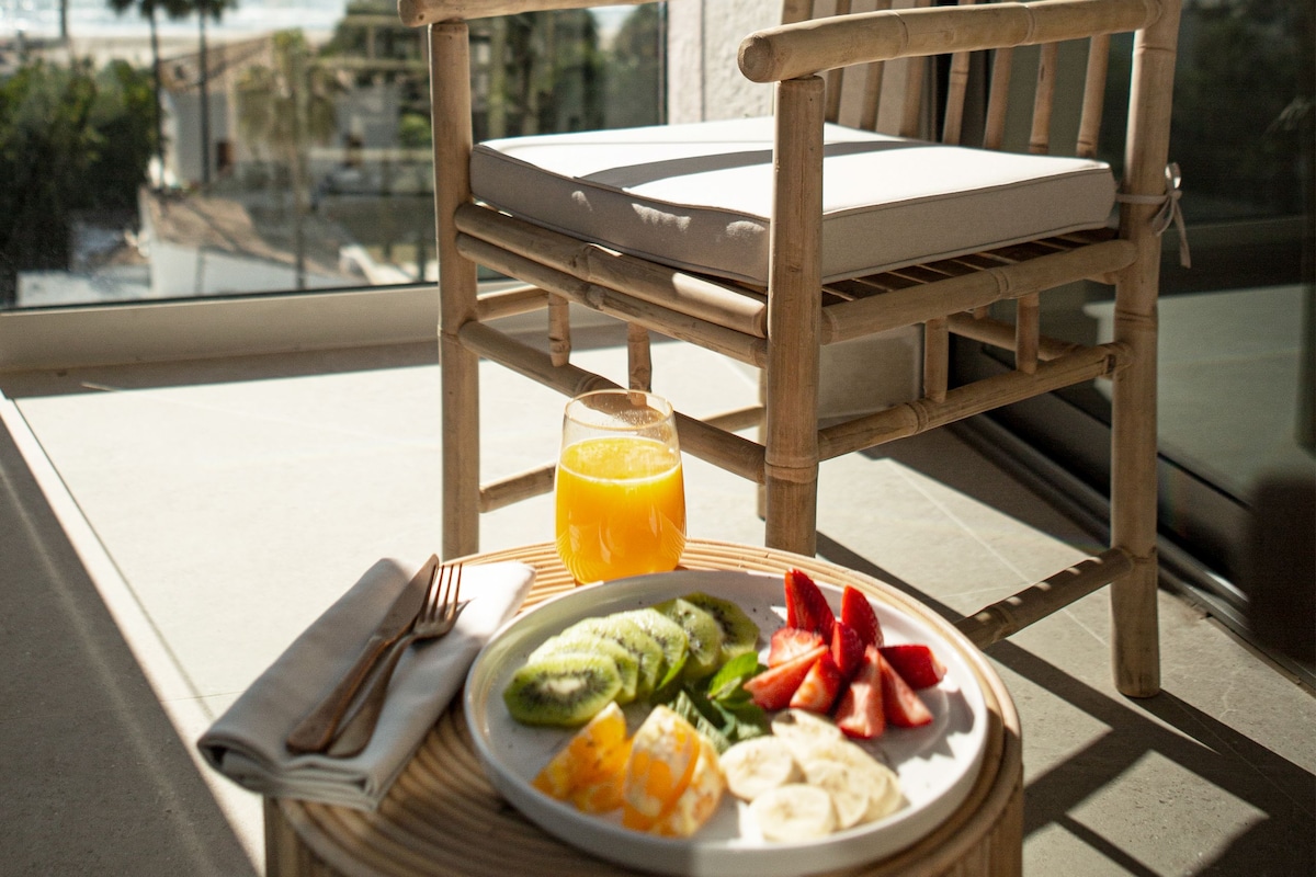 A wooden chair with a cushion on a balcony with a table, a plate of fruit, and a glass of orange juice.