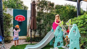 Kinderen spelen op een glijbaan en klimrek in een achtertuin met een basketbalveld en een groene muur.