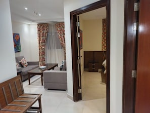 A living room with a sofa, a coffee table, and a wooden bench. There is a door leading to another room. - Golden New Heraa (Jeddah)