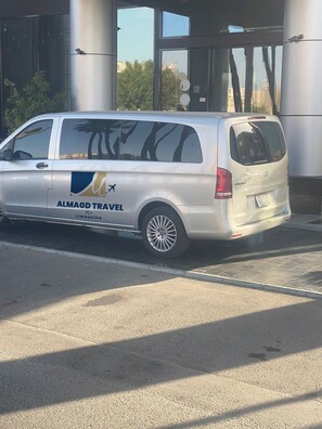 Een zilveren busje met de tekst 'ALMAGD TRAVEL' en 'Limousine' staat geparkeerd voor een gebouw met grote ramen en een metalen constructie.