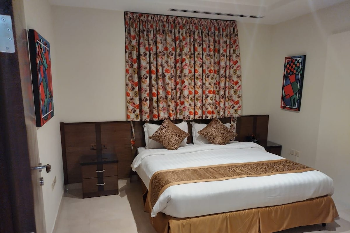 A bedroom with a bed, wooden headboard, curtains, and two paintings on the walls.