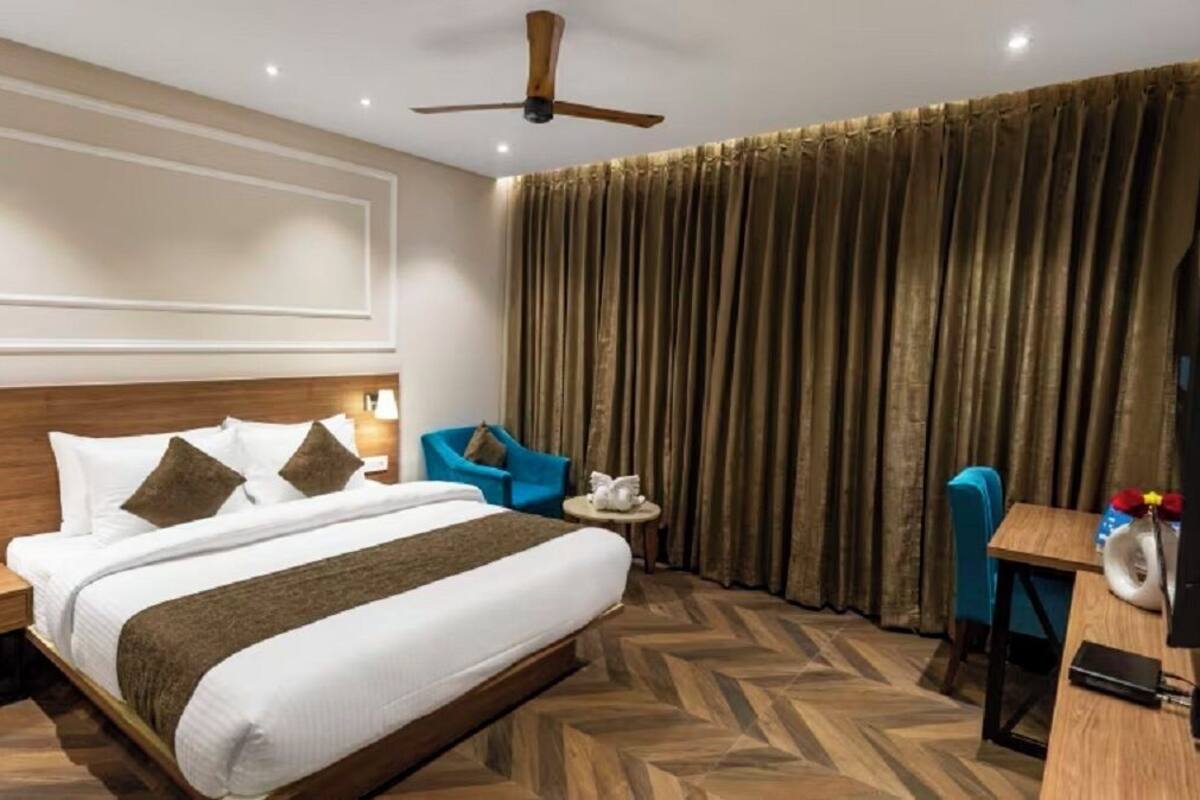 Habitación de hotel con cama, escritorio, silla, ventilador de techo y ventana grande con cortinas.