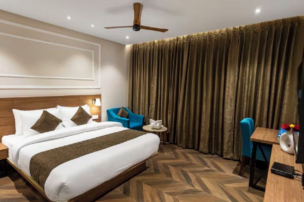 Een hotelkamer met een bed, een bureau, een stoel, een plafondventilator en een groot raam met gordijnen.