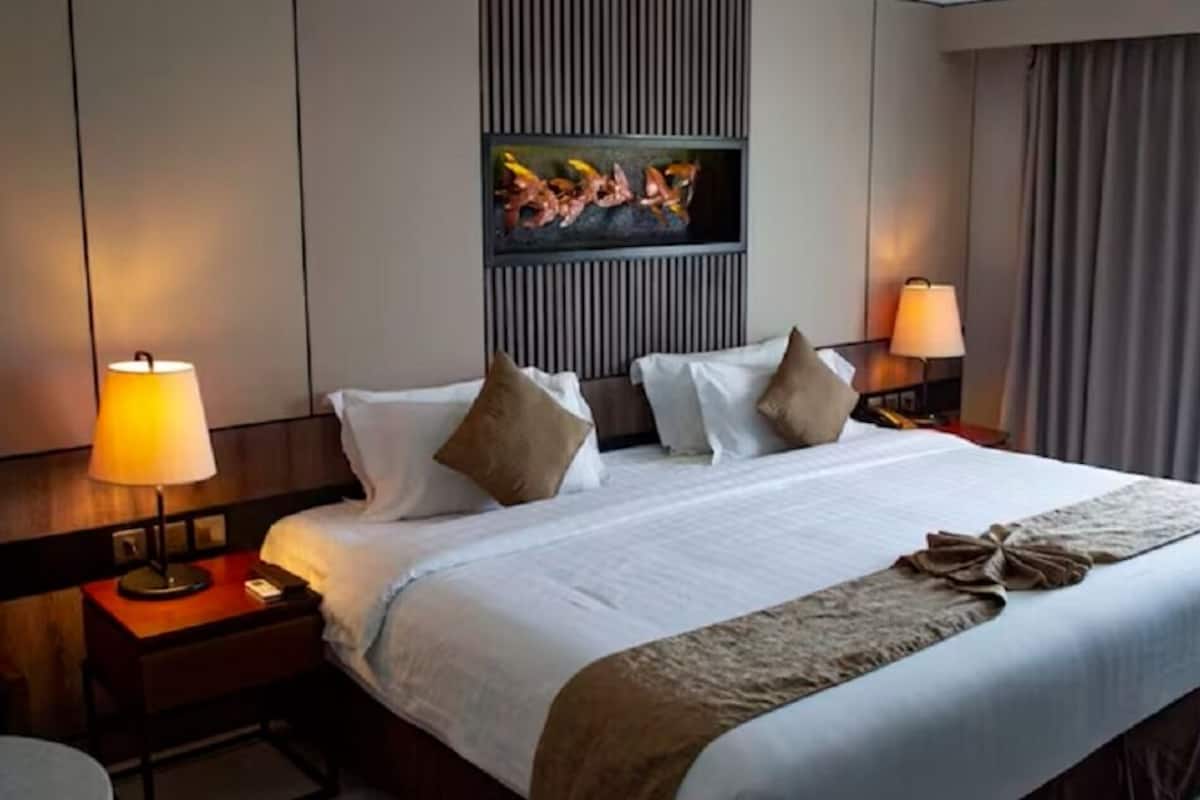 Ein Hotelzimmer mit einem großen Bett, zwei Nachttischlampen, einem flachbildfernseher an der Wand und einem Fenster mit Vorhängen.