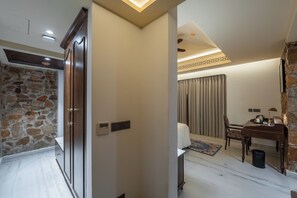 A hotel room with a stone wall, wooden wardrobe, a bed with curtains, a desk with a lamp, and a small trash bin. - Juna Mahal Jungle Resort (Sawai Madhopur)