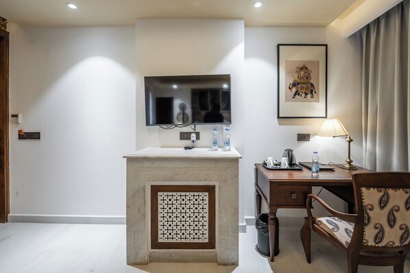 A hotel room with a marble bathroom vanity, a mounted flat-screen TV, a framed artwork above the TV, a desk with a lamp, and a patterned chair. - Juna Mahal Jungle Resort (Sawai Madhopur)