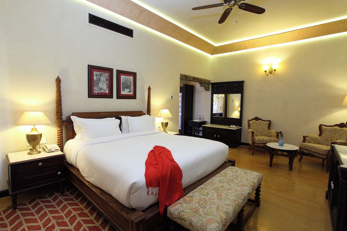 A hotel room with a large bed, bedside lamps, a wooden headboard, a bench, a small round table, two chairs, a mirror, and a ceiling fan.