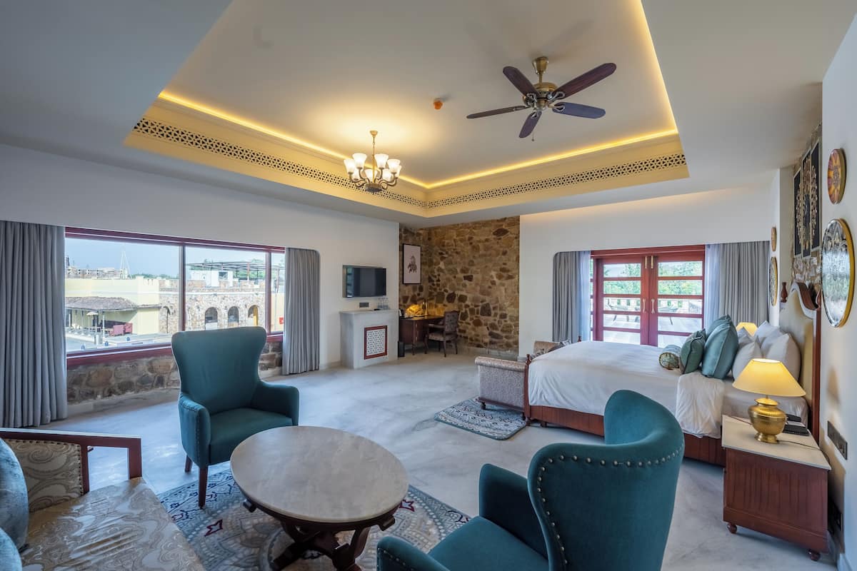 A spacious room with a large bed, a desk, and a chair. There is a television, a chandelier, and a ceiling fan.