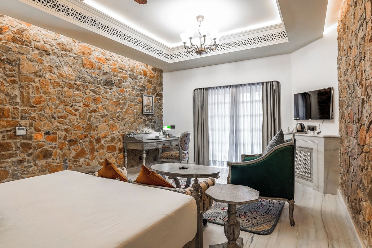 A hotel room with a stone wall, a bed, a desk with a chair, a television, and a window with curtains.