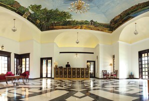 A lobby with a reception desk, seating area, chandeliers, and a mural on the ceiling.