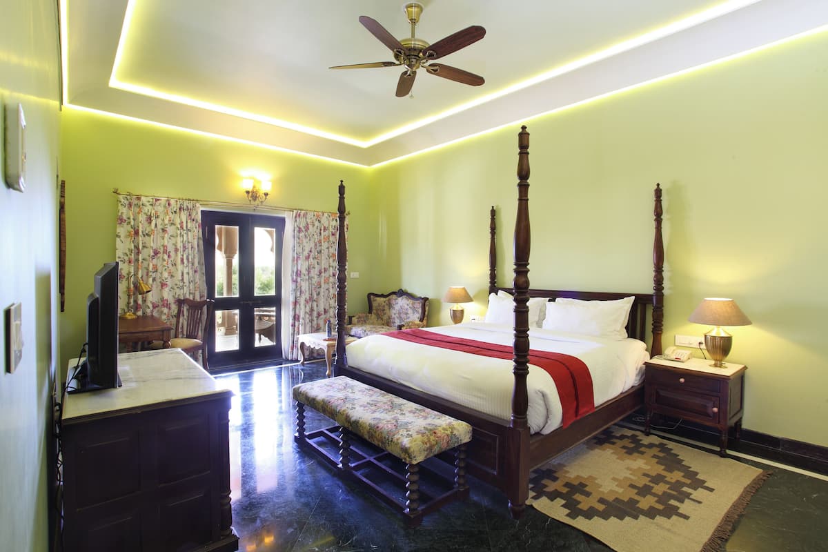 A four-poster bed with a wooden headboard, a bench with floral upholstery, a bedside table with a lamp, and a ceiling fan.