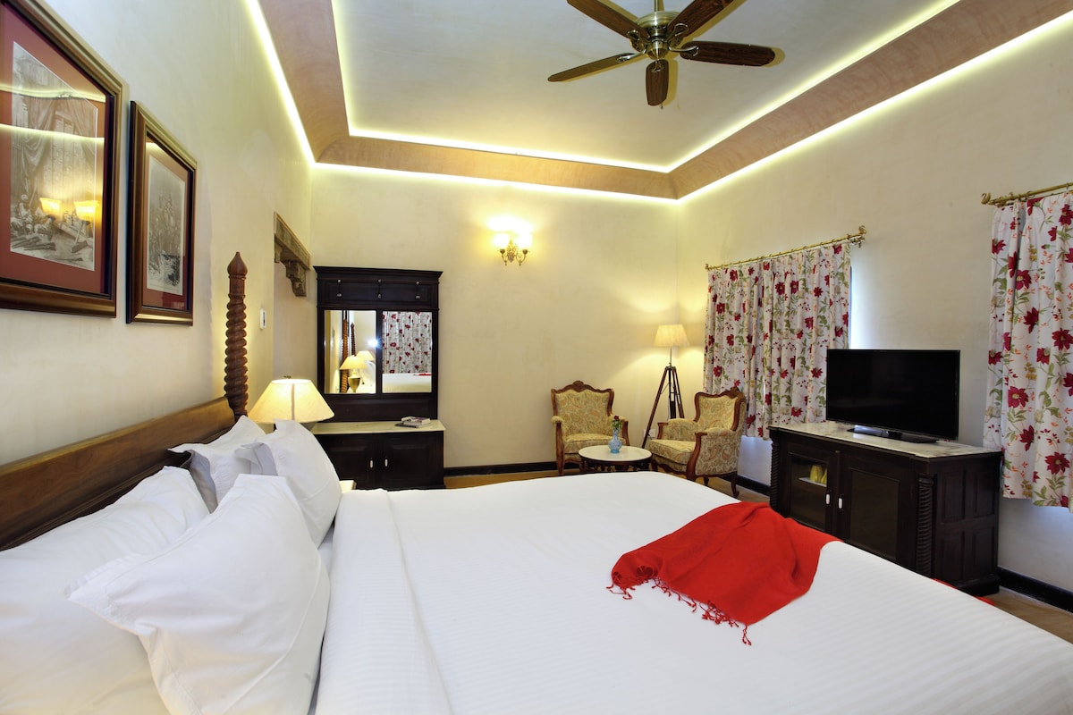 A hotel room with a bed, a television, a ceiling fan, a desk with a lamp, and a chair.