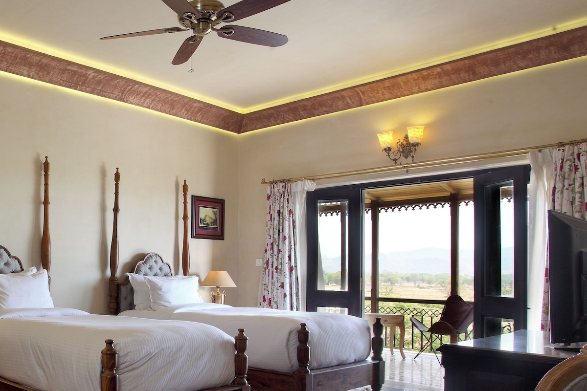 A bedroom with two beds, a ceiling fan, a desk with a chair, and a view through an open door.