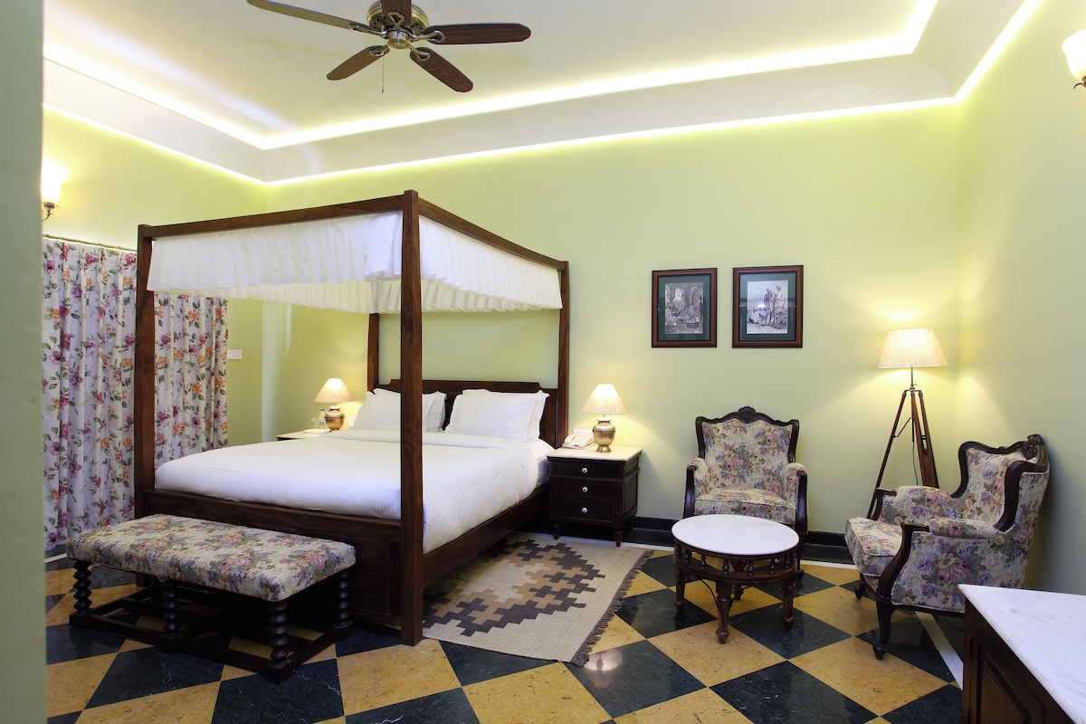 A four-poster bed with a canopy, a bench, two chairs, a small table, and a floor lamp in a room with a checkered floor and a ceiling fan.