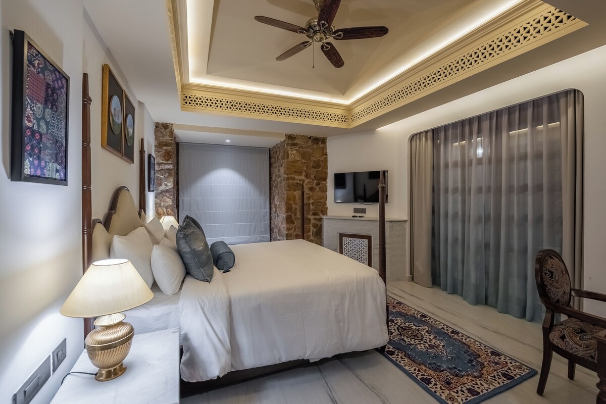 A hotel room with a bed, a TV, a ceiling fan, a stone accent wall, and a large window with curtains.