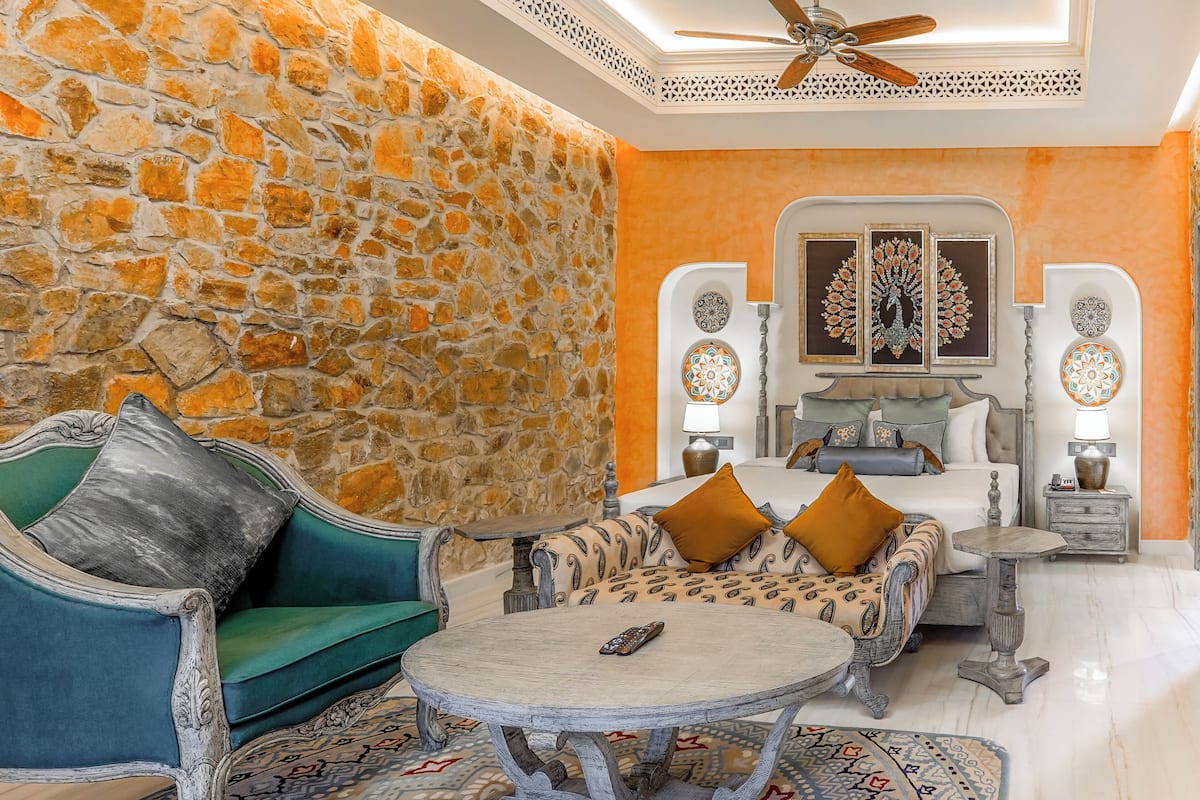 A room with a stone wall, a bed with patterned bedding, a green sofa, a round table, and two wall-mounted lamps.