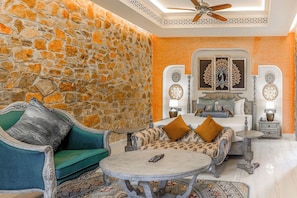 Una habitación con pared de piedra, una cama con ropa de cama estampada, un sofá verde, una mesa redonda y dos lámparas fijadas a la pared.