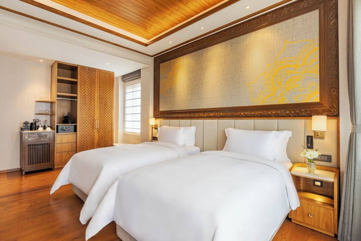 Habitación de hotel con dos camas, un armario de madera, una obra de arte grande en la pared y una mesita de noche con lámpara.