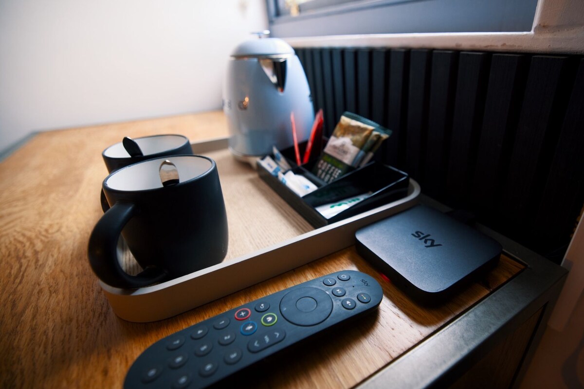 A wooden table with a remote control, a black mug, a kettle, and a Sky storage box.