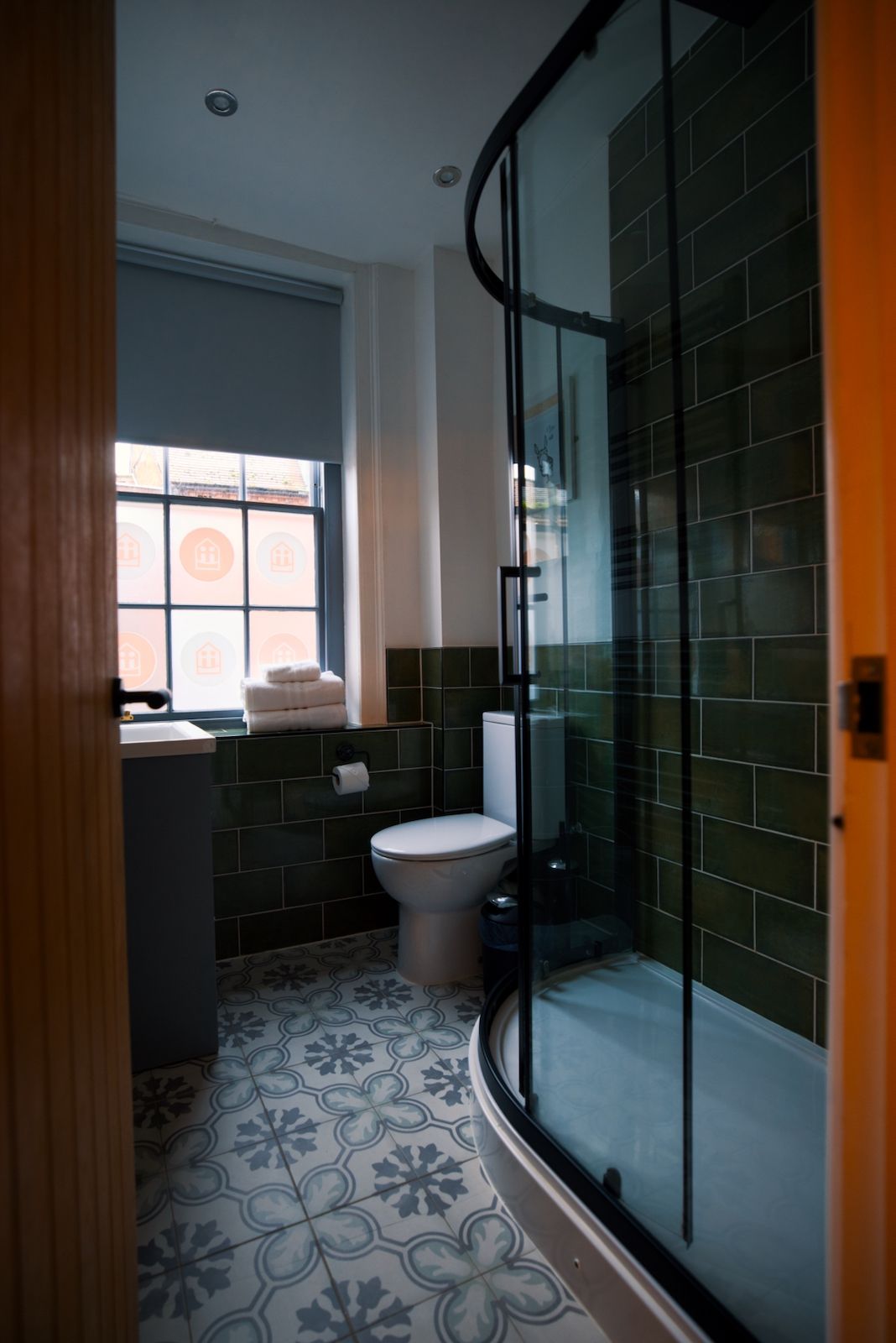 A bathroom with a toilet, a glass shower enclosure, a window with a view, and patterned floor tiles.