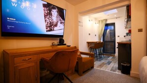 A hotel room with a wooden desk, a brown chair, a TV displaying a movie, a small kitchen area, and a window with curtains. - City Rooms by Hiive (Worcester)