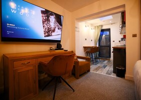 Habitación de hotel con un escritorio de madera, una silla marrón, un televisor mostrando una película, una pequeña cocina y una ventana con cortinas.