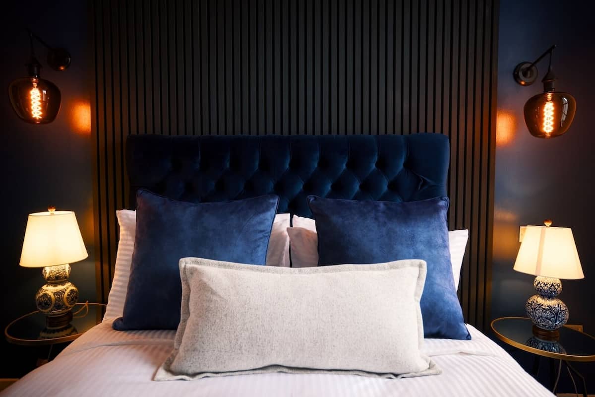 A bed with a tufted headboard, two blue pillows, a white pillow, and two bedside lamps with patterned bases.